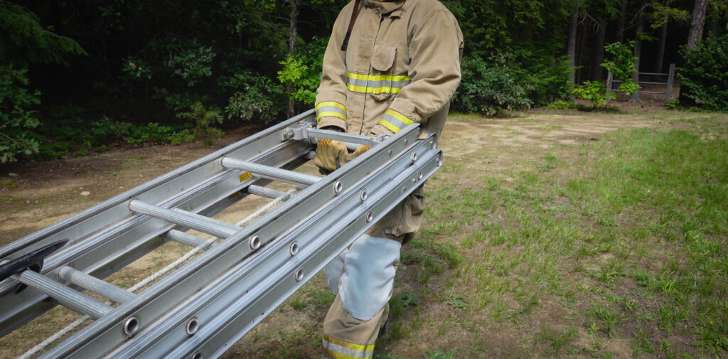A firefighter nests a roof ladder on top of an extension ladder, places hand tools on top, lifts one end, and drags the collected items along the ground.