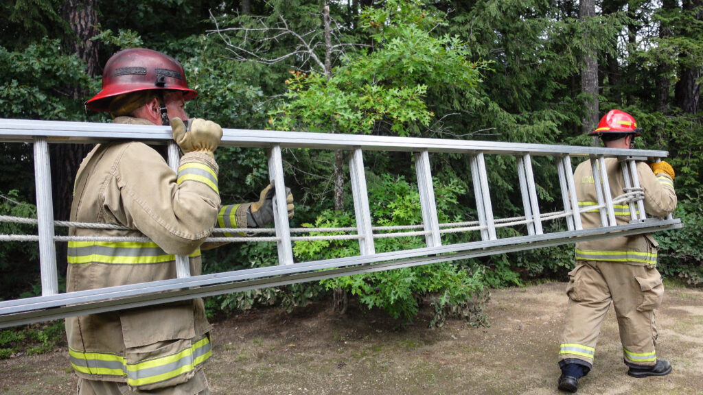 Spaced along the length of an extension ladder, two firefighters carry it by inserting their arms between ladder rungs and walk together.
