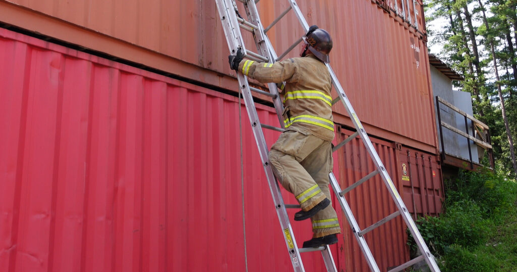 A firefighter climbing an extension ladder with a roof ladder slung over his shoulder.